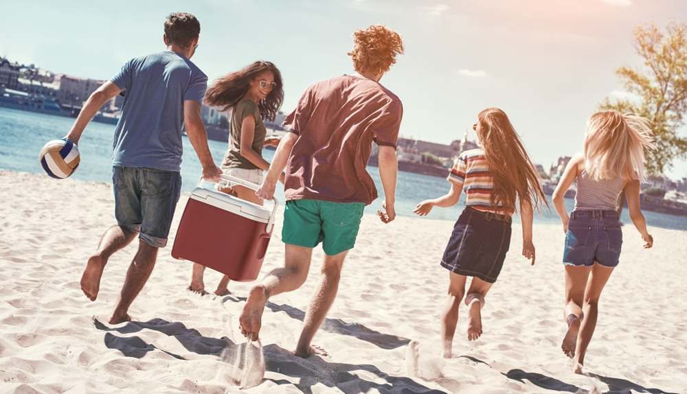 Friends on the beach. Rear view of cheerful young people walking by the beach to the sea while two men carrying plastic cooler